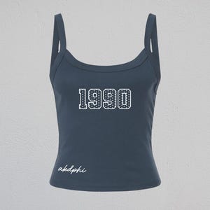 May include: A dark blue tank top with a scoop neckline and thick shoulder straps. The center of the chest features the number 1990 in a white, dotted varsity-style font. The lower left side displays the text akdphi in a white cursive script.