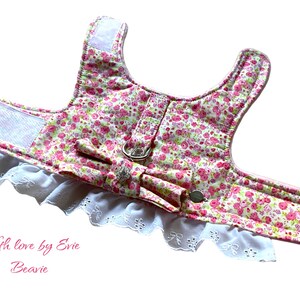 Può includere: Un'imbracatura per cani floreale rosa e bianca con un bordo in pizzo bianco e un fiocco rosa. L'imbracatura ha un pannello in rete bianca sul retro e una fibbia argentata. Il testo "With love by Evie Beavie" è stampato nella parte inferiore dell'immagine.