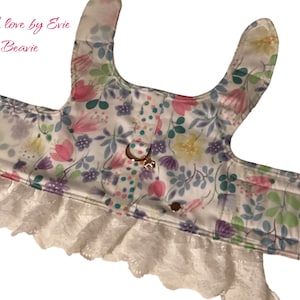 Puede incluir: Un arnés blanco para perro con estampado floral y ribete de encaje blanco. El arnés tiene dos correas que van sobre los hombros del perro y una correa que va alrededor del pecho del perro. El arnés está decorado con un estampado floral en tonos rosa, morado, amarillo y verde. El arnés tiene un ribete de encaje blanco alrededor del borde inferior.