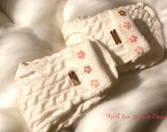 May include: Two white knitted baby booties with pink flower embellishments and a brown tag that says "With love by Blue Bear".