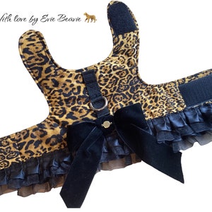 Pode incluir: Arnês para cães com estampa de leopardo, laço de veludo preto e acabamento com babados pretos. Possui alças ajustáveis e um anel em D para a trela. O texto "With love by Evie Beavie" é visível.