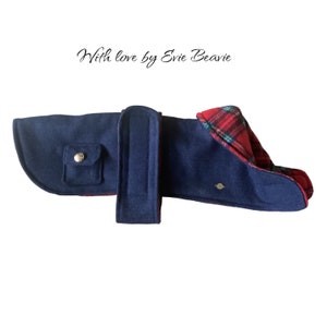 May include: A navy blue dog coat with a red and green tartan lining. The coat has a pocket on the front and a strap around the neck. "With love by Evie Beavie" is written on a white background above the coat.