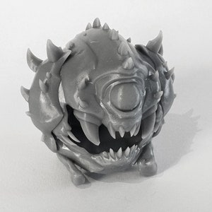 Op de afbeelding: Een grijs 3D-geprint miniatuur van een wezen met een enkel groot oog, scherpe tanden en stekels. Het wezen is rond en heeft een dreigende uitdrukking.