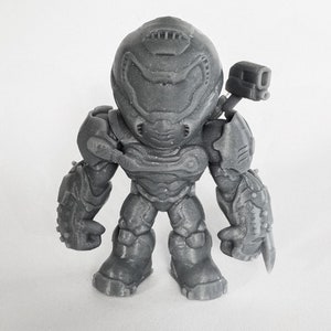 Puede incluir: Una figura de plástico gris y detallada de un personaje de videojuego. El personaje lleva un casco y una armadura, y sostiene un arma.