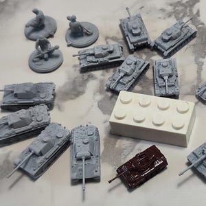 Könnte beinhalten: Eine Ansammlung von Miniatur-Spielzeugpanzern und Soldaten in Grau ist auf einer Oberfläche mit Marmormuster angeordnet. Ein weißer Lego-Stein und ein dunkelbrauner Panzer sind ebenfalls vorhanden. Die Panzer variieren im Design, was auf ein militärisches Thema hindeutet.