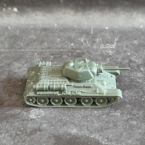 May include: A miniature olive-green toy tank, featuring a detailed design with a rotating turret and gun barrel. The tank's treads are visible. The model is likely plastic and rests on a grey surface. A classic toy.