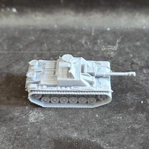 Puede incluir: Un peque&ntilde;o modelo de tanque gris claro con un ca&ntilde;&oacute;n largo y orugas detalladas. El veh&iacute;culo militar en miniatura se encuentra sobre una superficie oscura y texturizada.