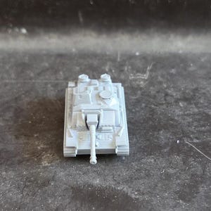 Puede incluir: Un peque&ntilde;o tanque de juguete gris claro con un ca&ntilde;&oacute;n largo y detalles. El modelo en miniatura tiene un cuerpo rectangular, dos torretas y orugas. El tanque est&aacute; sobre un fondo oscuro y texturizado.