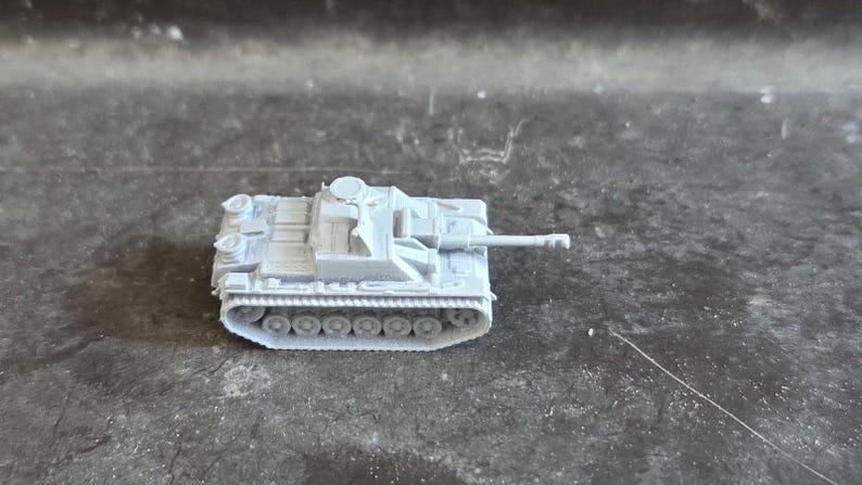 Puede incluir: Un peque&ntilde;o modelo de tanque gris claro con un ca&ntilde;&oacute;n largo y orugas detalladas. El veh&iacute;culo militar en miniatura se encuentra sobre una superficie oscura y texturizada.