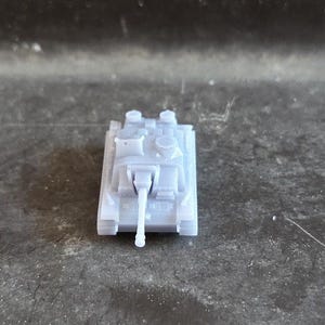 Puede incluir: Un peque&ntilde;o modelo de tanque gris claro, visto desde arriba, con un ca&ntilde;&oacute;n largo y detalles precisos. El tanque se encuentra sobre un fondo oscuro y texturizado.