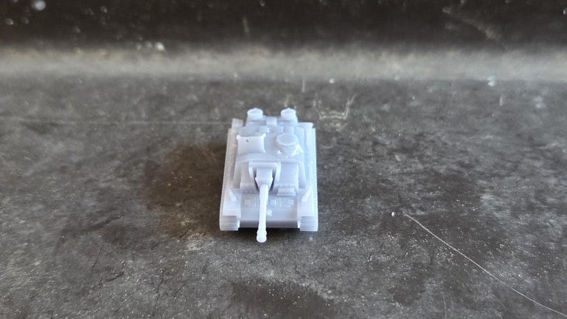 Puede incluir: Un peque&ntilde;o modelo de tanque gris claro, visto desde arriba, con un ca&ntilde;&oacute;n largo y detalles precisos. El tanque se encuentra sobre un fondo oscuro y texturizado.