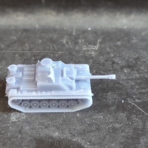 Puede incluir: Un peque&ntilde;o modelo de tanque gris claro, con un ca&ntilde;&oacute;n largo y orugas detalladas. El veh&iacute;culo en miniatura se encuentra sobre una superficie oscura y texturizada, mostrando su intrincado dise&ntilde;o. Este modelo es probablemente un objeto de colecci&oacute;n o para un diorama.