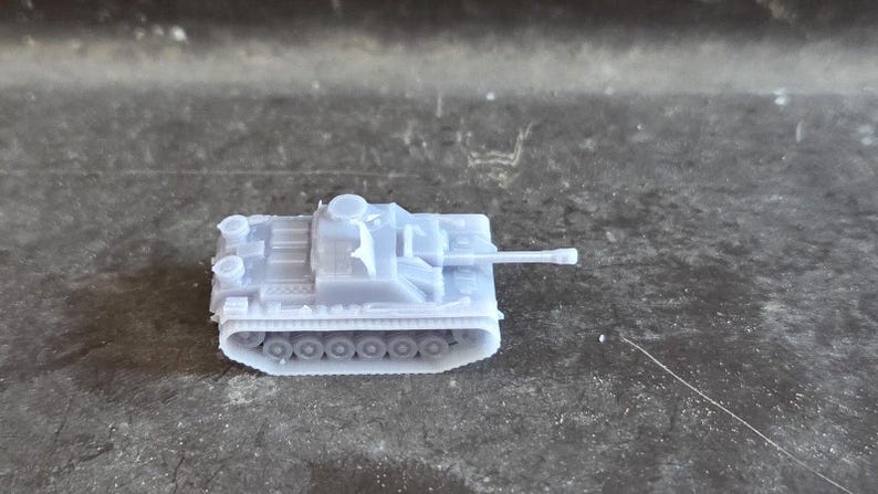 Puede incluir: Un peque&ntilde;o modelo de tanque gris claro, con un ca&ntilde;&oacute;n largo y orugas detalladas. El veh&iacute;culo en miniatura se encuentra sobre una superficie oscura y texturizada, mostrando su intrincado dise&ntilde;o. Este modelo es probablemente un objeto de colecci&oacute;n o para un diorama.