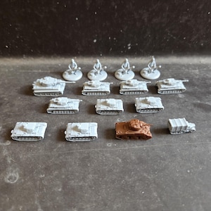 Op de afbeelding: Een verzameling miniatuur militaire voertuigen en figuren, waaronder elf kleine grijze tanks, één bruine tank, één kleine grijze vrachtwagen en vier kleine grijze soldaatfiguren, allemaal in rijen op een donker oppervlak geplaatst.