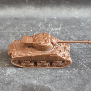 Op de afbeelding: Een kleine, bruine speelgoedtank met een lange loop en gedetailleerde rupsbanden. Het miniatuur militaire voertuig is gemaakt van plastic en staat op een grijze ondergrond. De tank is een replica van een klassiek militair voertuig.