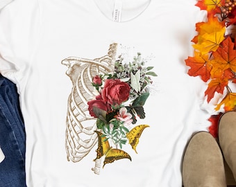 May include: White t-shirt featuring a gold-toned rib cage design with red roses, butterflies, and greenery. The graphic tee has a crew neck and short sleeves. The shirt is made of a soft, comfortable material.