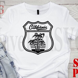 May include: White t-shirt with a black graphic of a California road sign with palm trees and the text "California".