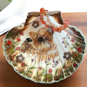 May include: A decorative seashell ring dish with a painted floral design and the words "Rings & Things." The dish holds several rings and a beaded necklace. The rim is trimmed with a green and gold border.