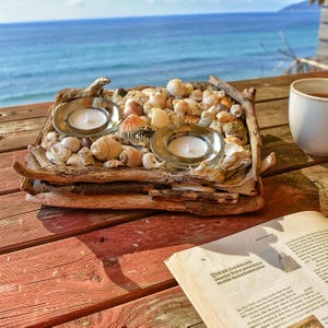 Könnte beinhalten: Ein rechteckiger Kerzenhalter aus Treibholz, gefüllt mit Muscheln und zwei Glashaltern mit brennenden weißen Kerzen. Der Halter steht auf einem roten Holztisch mit Blick auf das Meer und einer Tasse Tee.