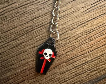 May include: A black coffin-shaped keychain charm with a white skull and red accents. The charm is attached to a silver chain and keyring. The keychain is on a wooden surface.