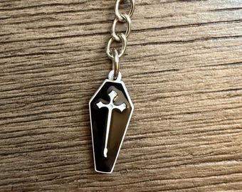 May include: A silver keychain with a black coffin-shaped charm. The charm features a white cross design. The keychain is attached to a silver chain and ring, set against a wooden background.