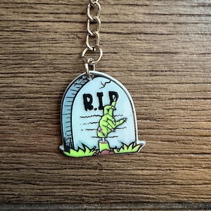 May include: A Halloween-themed keyring with a tombstone design. The tombstone is light blue with the letters "R.I.P." in black. A green zombie hand emerges from the ground. The keyring is attached to a silver chain.