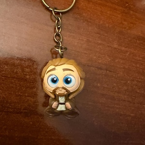 Puede incluir: Un llavero con una figura de dibujos animados con cabello castaño claro, barba y ojos azules grandes. La figura lleva un atuendo marrón y está unida a un llavero y cadena dorados. El fondo es una superficie de madera marrón oscuro.