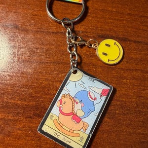 Pode incluir: Um chaveiro com argola e corrente prateadas, com um pingente de carinha sorridente amarela e um pingente de carta de tarô. A carta de tarô retrata um cavalo de balanço e um personagem azul com uma pipa, rotulado como "The Sun".