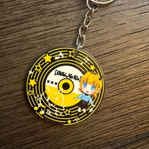 Może przedstawiać: Okrągły, żółto-czarny brelok do kluczy z postacią z kreskówki i napisem "[Oshi No Ko]". Brelok ma srebrny łańcuszek i kółko. Projekt zawiera nuty i gwiazdy.