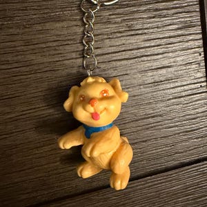 May include: A tan-coloured dog keyring with a blue collar and a pink tongue sticking out. The dog is attached to a silver chain and keyring. The keychain is made of plastic and has a cartoonish design.