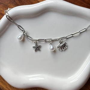 Könnte beinhalten: Eine silberne Kette mit drei Anhängern: ein Seestern, eine Muschel und eine Kunstperle. Die Kette wird auf einem weißen Teller mit gewelltem Rand präsentiert, der auf einer warmen Holzoberfläche liegt.
