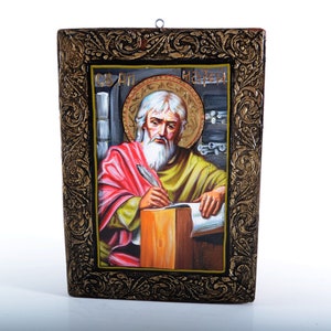 Puede incluir: Un icono enmarcado que representa a un hombre barbudo con halo, sosteniendo una pluma y escribiendo en un libro. El icono está pintado en un estilo tradicional con detalles dorados y un marco negro.