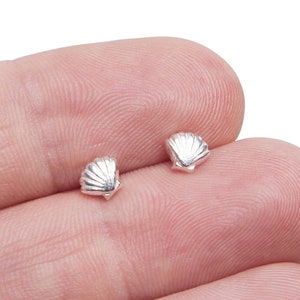 May include: Two small, silver seashell-shaped stud earrings. The earrings have detailed ridges, replicating the texture of a real seashell. The earrings are displayed against a neutral background.