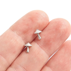 Puede incluir: Dos pequeños pendientes plateados con forma de seta. Los pendientes se sostienen en la mano de una persona sobre un fondo blanco. Los sombreros de las setas son lisos y los tallos tienen un aspecto texturizado. Un accesorio caprichoso.