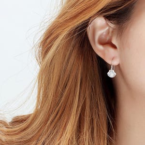May include: A silver seashell earring hangs from a person's ear. The earring is a small, detailed replica of a seashell, with a curved, ridged design. The earring is attached to a simple hook.