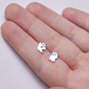 Könnte beinhalten: Ein Paar silberne Ohrstecker in Geisterform. Jeder Ohrring hat ein einfaches Geisterdesign mit ausgeschnittenen Augen und Mund. Die Ohrringe sind klein und zart, perfekt für den Alltag.