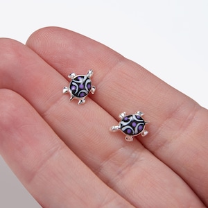 Puede incluir: Un par de pendientes de plata con forma de tortuga con caparazones con patrones en blanco y negro y detalles en morado.