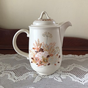 Peut inclure: Une cafetière en céramique blanche avec un motif floral composé de fleurs brunes, orange et bleues. La cafetière a un couvercle avec un bouton sur le dessus.