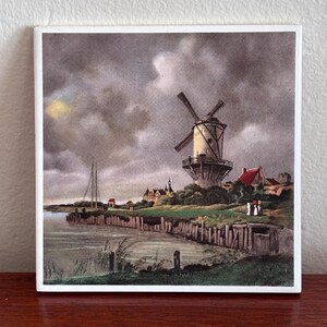 May include: Square ceramic tile with a vintage landscape scene. The artwork shows a Dutch windmill on a riverbank under a cloudy sky. Small buildings and figures are also visible, creating a picturesque scene.