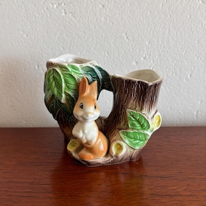 May include: A ceramic planter featuring a brown and orange bunny nestled in a tree trunk. The planter has a hollowed-out interior for plants, with green leaves and yellow accents. The bunny is detailed with a white belly and a sweet expression.