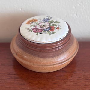 May include: A round wooden container with a floral ceramic lid. The lid has a white background with a colourful floral design in red, orange, blue, and purple. The container has a warm, natural wood grain finish.