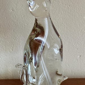 May include: A clear glass cat figurine with a rounded body, pointed ears, and a curled tail. The glass has a swirled, marbled effect with white and brown tones. The cat is sitting upright.