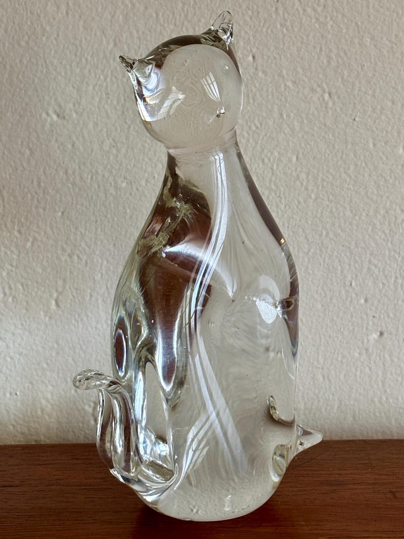 May include: A clear glass cat figurine with a rounded body, pointed ears, and a curled tail. The glass has a swirled, marbled effect with white and brown tones. The cat is sitting upright.