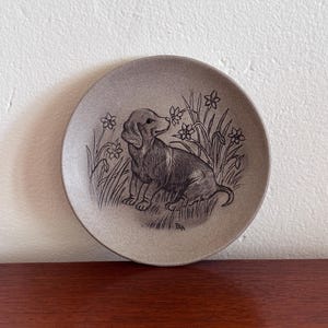 May include: A small, round, gray plate featuring a detailed illustration of a dachshund dog sitting in grass with flowers. The dog is in a seated position, with its head turned slightly. The plate has a textured surface.
