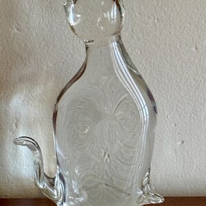 May include: A clear glass cat figurine with pointed ears, a curved tail, and a seated pose. The transparent sculpture has a smooth surface with internal swirls and bubbles, and sits on a wooden surface.