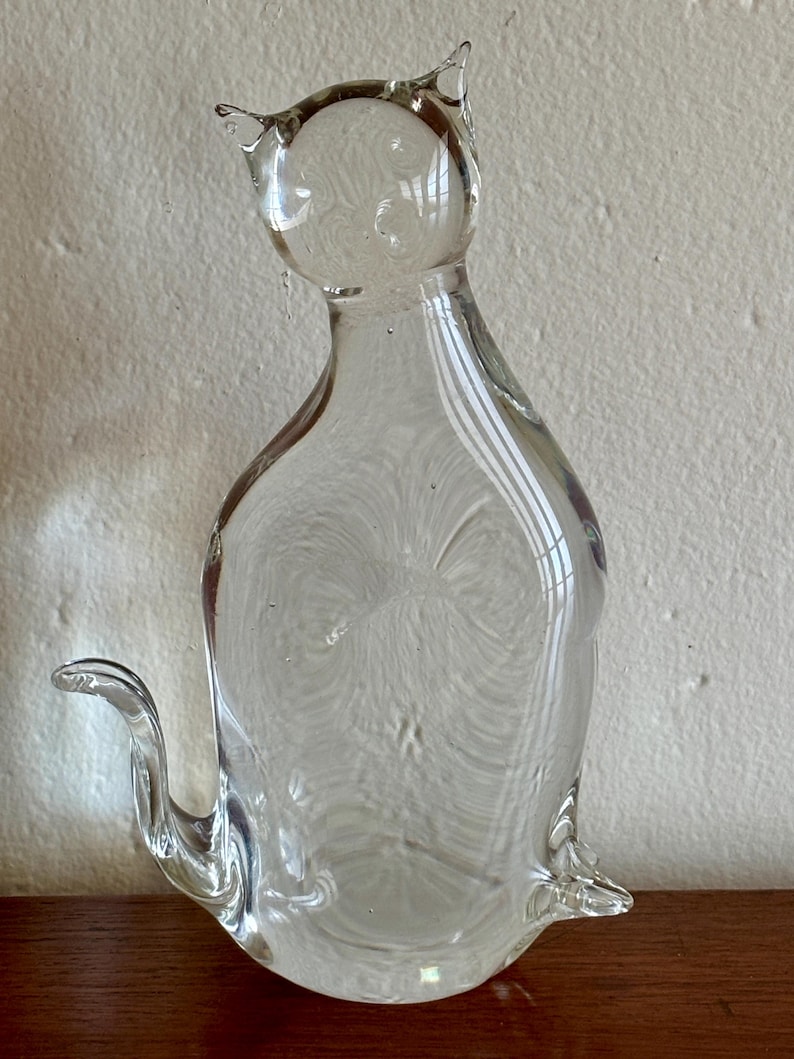 May include: A clear glass cat figurine with pointed ears, a curved tail, and a seated pose. The transparent sculpture has a smooth surface with internal swirls and bubbles, and sits on a wooden surface.