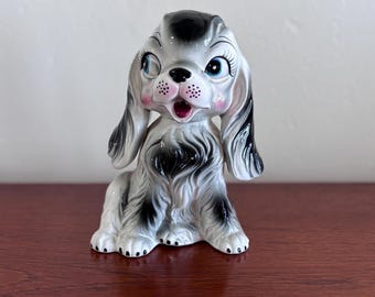May include: A vintage ceramic dog figurine in a seated position. The dog has a black and white coat, long floppy ears, and blue eyes. Its mouth is open, revealing a pink interior. The figurine has a glossy finish and is likely a decorative item.