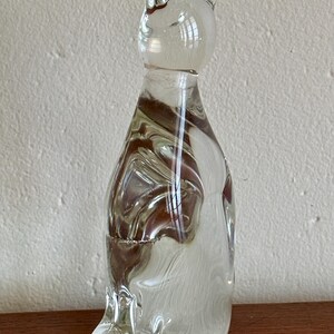 May include: A clear glass cat figurine with brown and white swirls throughout the body. The cat has pointed ears and a rounded head. The figurine is sitting on a wooden surface.