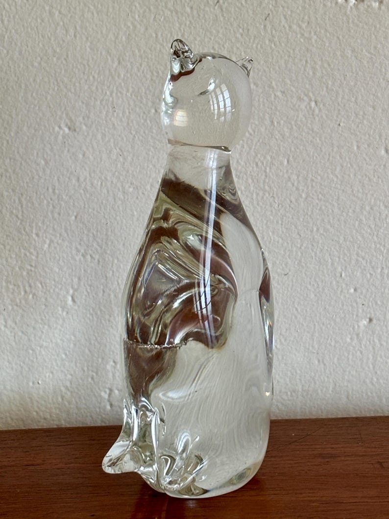 May include: A clear glass cat figurine with brown and white swirls throughout the body. The cat has pointed ears and a rounded head. The figurine is sitting on a wooden surface.