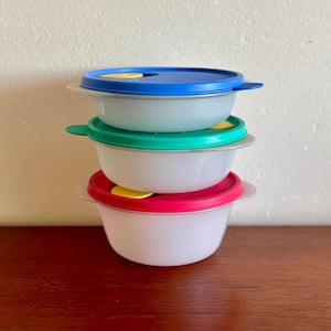 May include: Three plastic food storage bowls stacked on top of each other, each with a different coloured lid. The lids are blue, green, and red, with yellow tabs. The bowls are white with a silver rim.